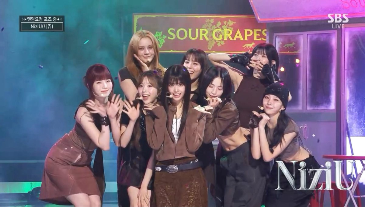 A group of female performers on stage pose together in brown outfits in front of a sign reading 'SOUR GRAPES', with on-screen text showing 'NiziU' and 'SBS LIVE'.