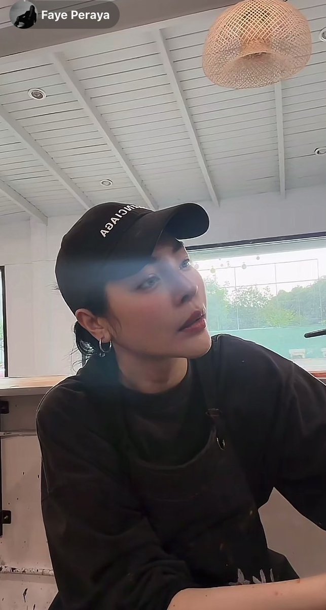 A person identified as 'Faye Peraya' wears a black shirt and a black 'MORISA' cap, looking upward and to the right in a room with a white slatted ceiling and a hanging rattan lamp.