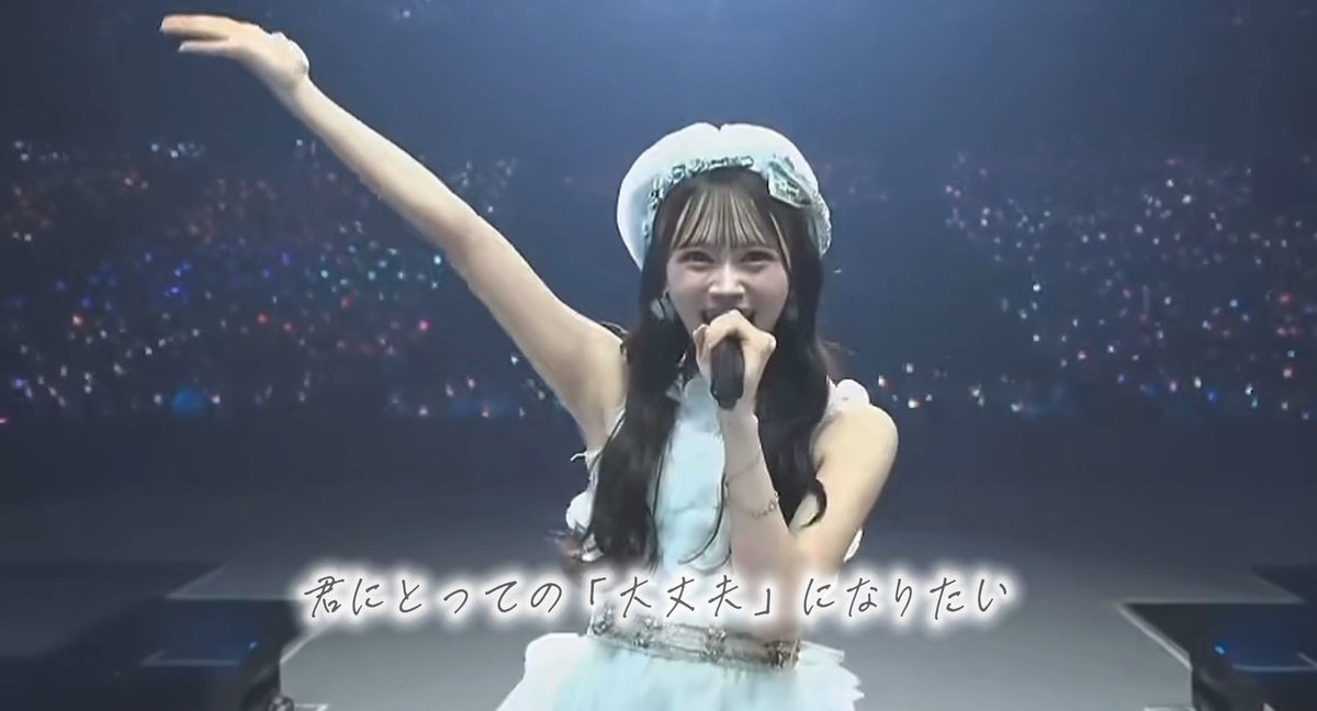 A young woman in a white dress and beret sings into a microphone on stage with Japanese text '君にとっての「大丈夫」になりたい' displayed.