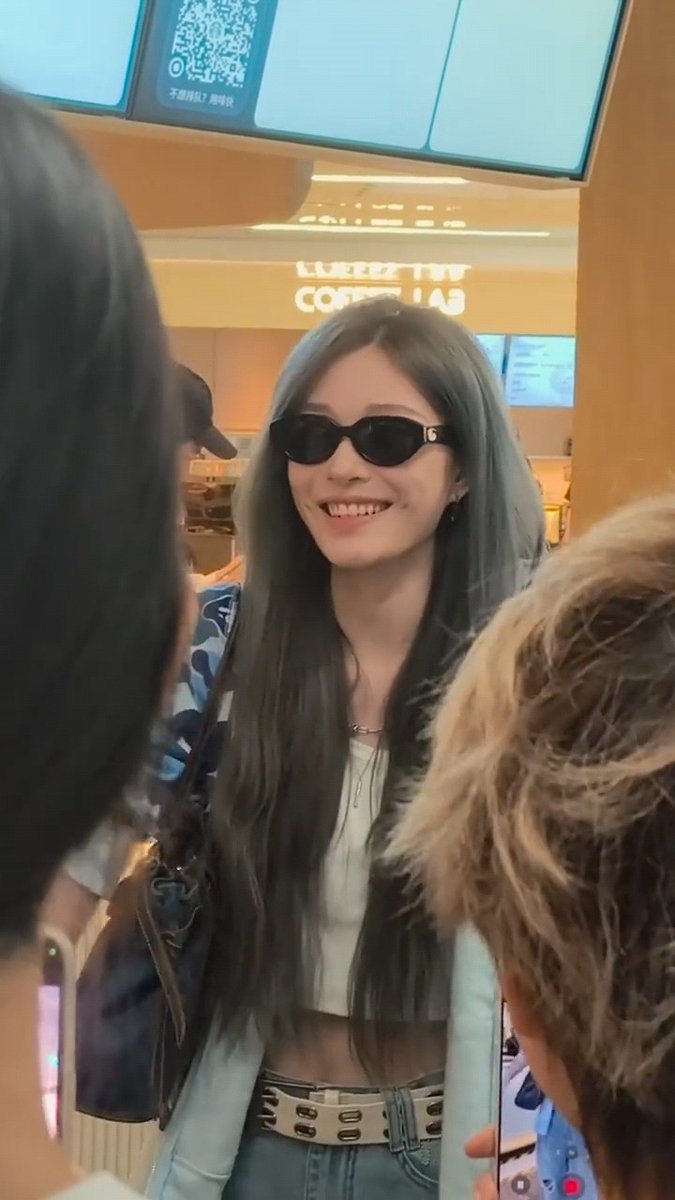 A young woman with long dark hair and bangs smiles while wearing dark sunglasses in a cafe setting.