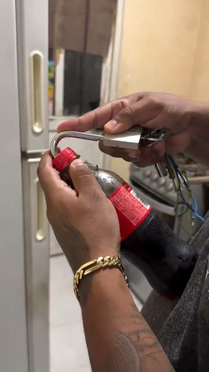 Close-up of a person's hands using a padlock on a large bottle, with one hand wearing a gold chain bracelet and a tattoo on the forearm.