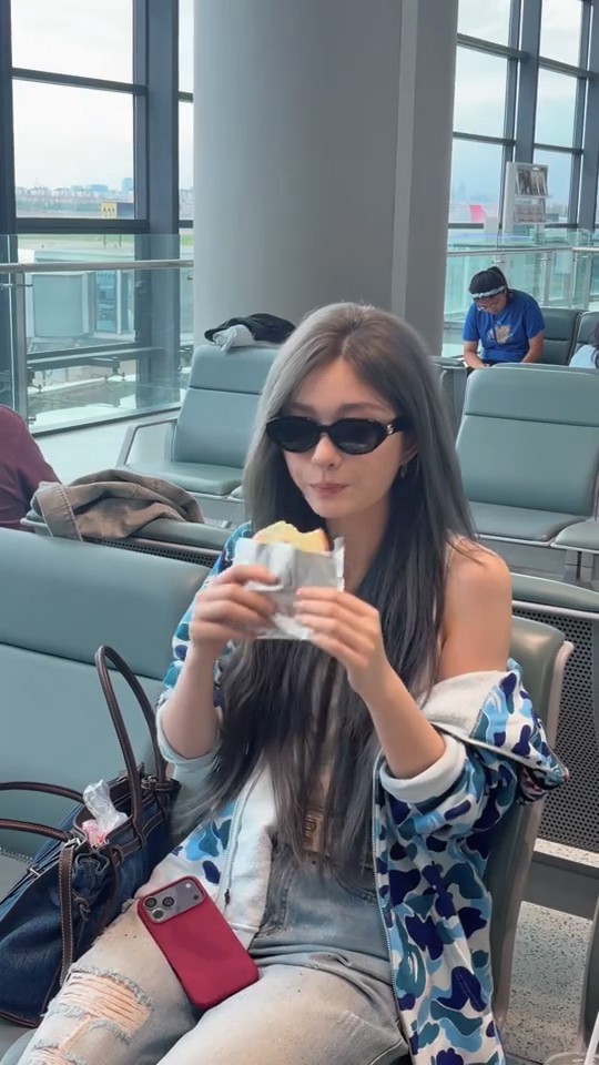 A young woman wearing sunglasses and a blue camouflage jacket sits in an airport waiting area, eating a snack wrapped in foil with a red smartphone resting on her lap.