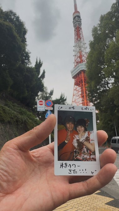 A hand holds a small Polaroid photo of two young people in front of Tokyo Tower, with Japanese handwriting on the white border.
