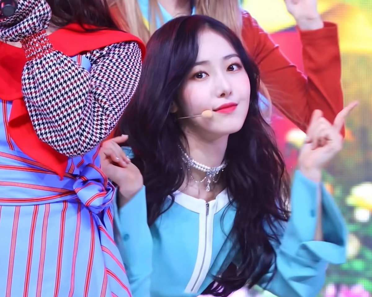 A K-pop idol with long dark hair in a light blue jacket making a heart shape with her fingers on stage.