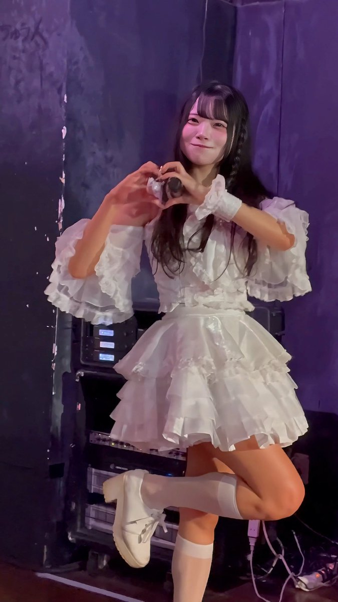 A young woman in a frilly white dress and knee-high socks poses backstage, making a heart shape with her hands while lifting one leg.