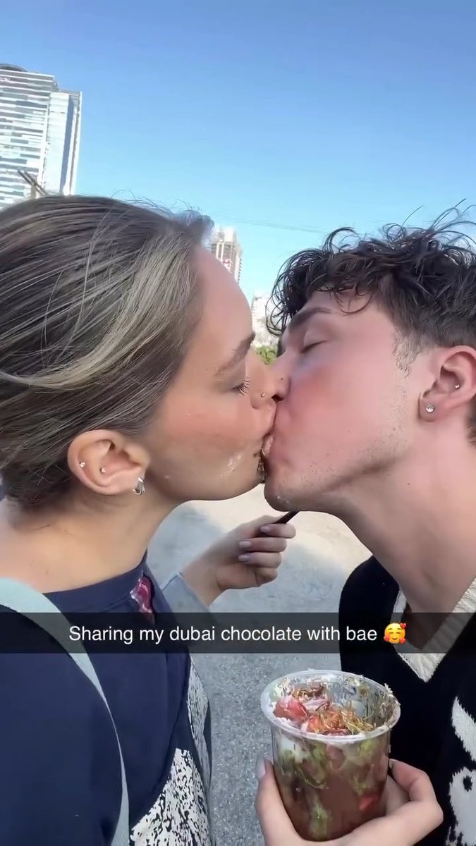 A couple shares a kiss outdoors while holding a cup of food, with text overlay reading 'Sharing my dubai chocolate with bae 🥰'.