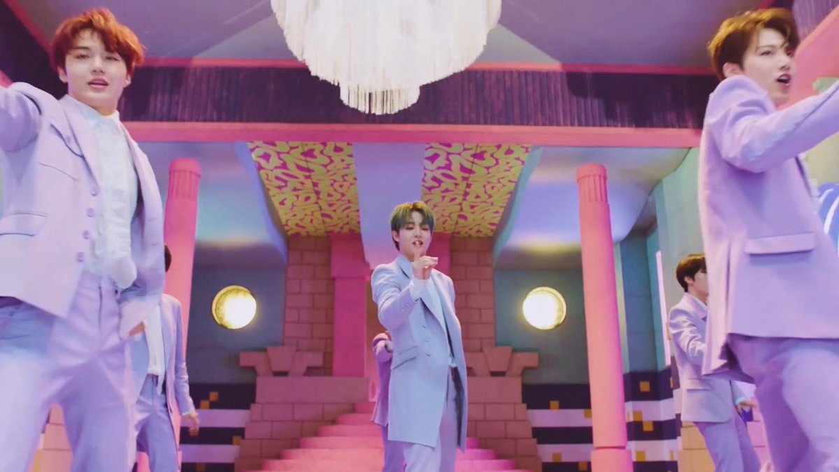 Several performers in matching lavender suits dance in a vibrantly colored pastel set featuring pink columns, a chandelier, and geometric ceiling patterns, suggesting a polished K-pop music video production. The coordinated styling and elaborate visual design hint at the choreographed aesthetic typical of a major label group, addressing the question of whether they belong to SM Entertainment.