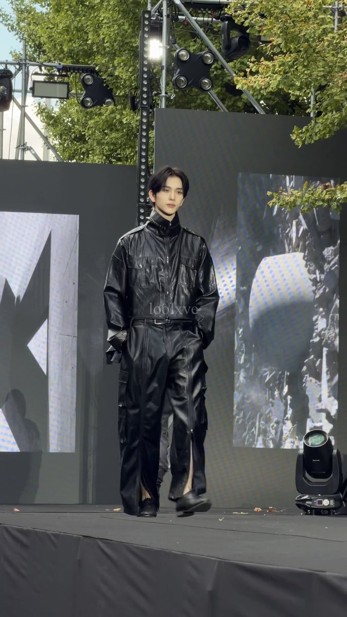 A young man with dark hair walks down an outdoor runway wearing an all-black leather outfit, with stage lights and trees visible in the background.