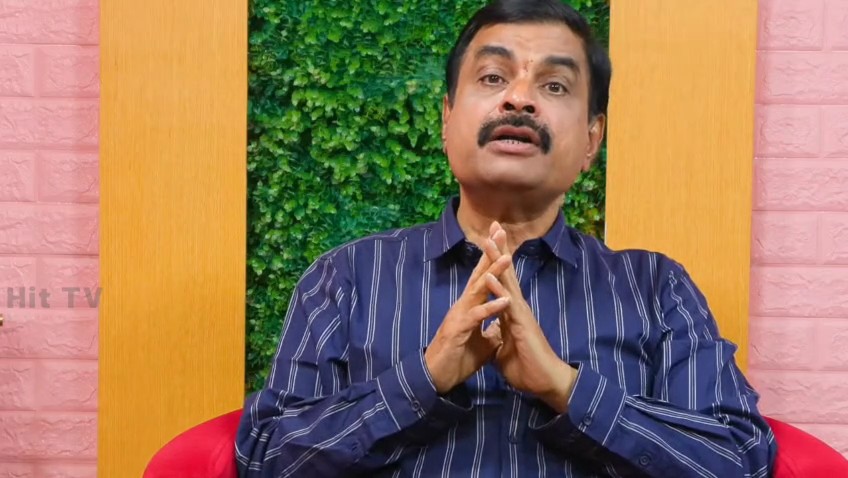 A man with a mustache wearing a blue striped shirt sits with hands clasped together, appearing to speak in front of a green plant wall with "Hit TV" watermark visible.