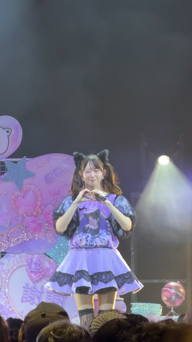 A performer on stage wearing a purple dress with a cat design, cat ear headband, and making a heart shape with her hands.