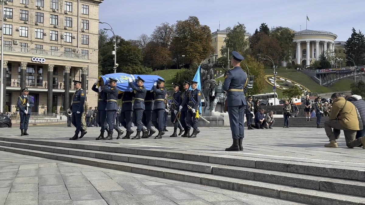 A group of uniformed military honor guards in dark blue dress uniforms carry a flag-draped coffin across a stone plaza in central Kyiv, with the Instytutska Street area visible behind them, while a photographer crouches in the foreground to document the funeral procession. This somber scene captures a fallen Ukrainian soldier being laid to rest, embodying the post's message that each casualty statistic represents a real person whose loss will be carried forever by grieving families.