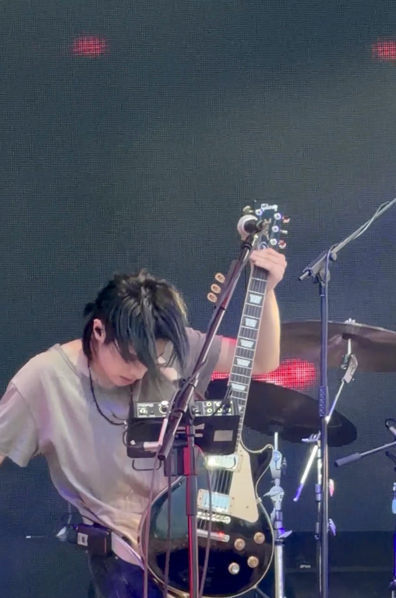 A young musician with dark hair performs on stage, head bowed over a black electric guitar, with drum equipment and red stage lights visible in the background.