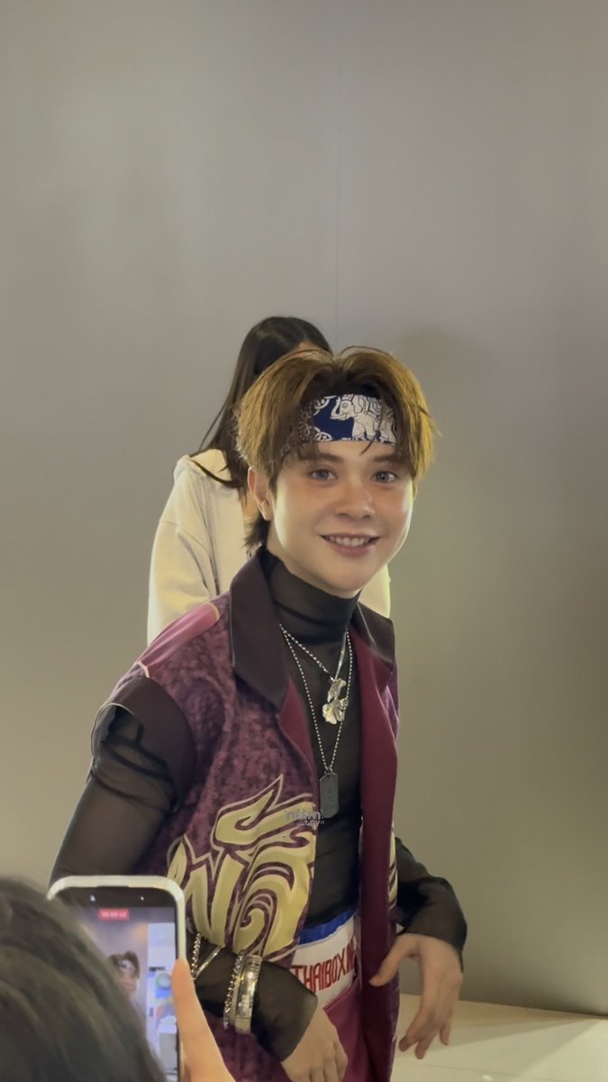 A young male idol wearing a blue bandana, a purple vest with gold designs, and multiple necklaces smiles at an event while a fan takes a photo in the foreground.