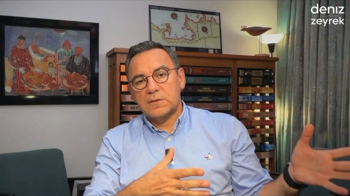 A man with glasses and a light blue shirt speaks in front of a bookshelf and a framed artwork; a watermark in the top-right corner reads "deniz zeyrek".