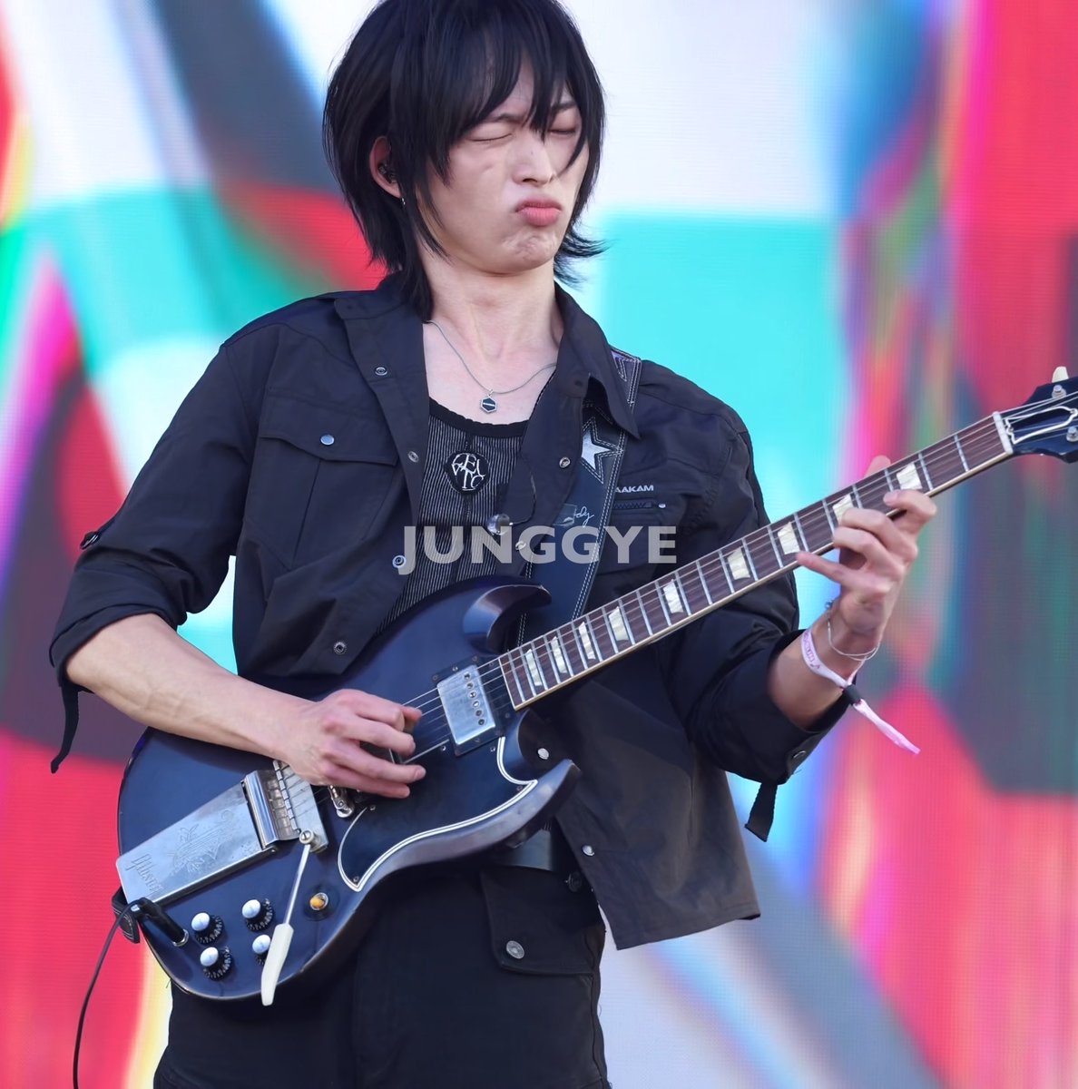 A guitarist with long black hair plays a Gibson electric guitar on stage, wearing a black jacket and a "JUNGGYE" watermark visible.