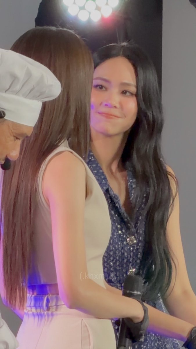 Two women interact on stage; one with long dark hair in a blue sequined outfit smiles warmly, while another in a white top stands nearby.