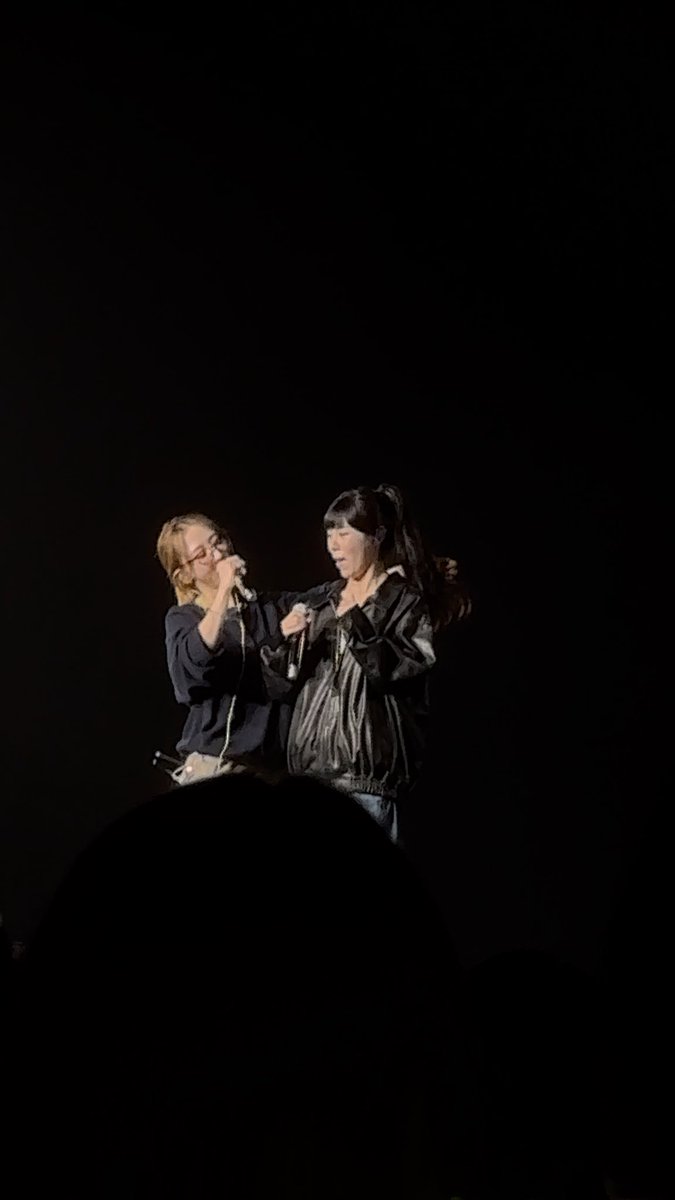 Two female performers sing on a dark stage, one wearing glasses and a dark jacket holding a microphone to her mouth, the other in a black leather jacket beside her.