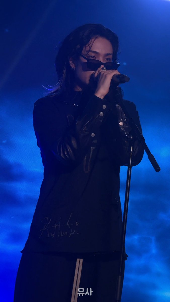 A performer with long dark hair sings into a microphone on stage under blue lighting, wearing a black leather jacket, dark sunglasses, and a black shirt with white script lettering. Korean text '육사' appears at the bottom.