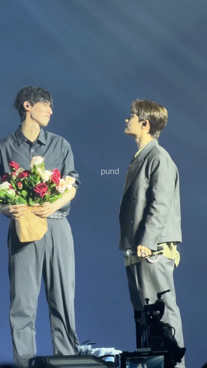 Two men share a tender moment on stage, with the taller one on the left holding a bouquet of pink and red roses while gazing affectionately at his shorter companion in a gray suit holding a microphone. The watermark "pund" appears on the image, and the caption "Wkwkwk gemes bangeetttt" (Indonesian for "cuteness overload, seriously!") reflects the heartwarming, fan-capturing interaction between the two performers.