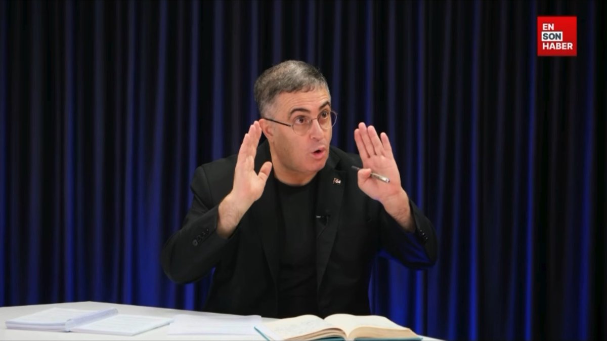 A man with glasses, wearing a black suit jacket over a black shirt, gestures emphatically with both hands raised while speaking in front of a dark blue curtain, with an open book and papers on the table before him. The "EN SON HABER" logo appears in the top right corner, and the image captures his animated expression matching his critical commentary about public authority's failure to oversee fund victims.