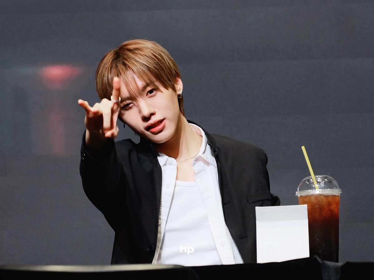 A young man with brown hair in a black blazer over a white shirt points toward the camera at what appears to be an event, with an iced drink and a white packet nearby.