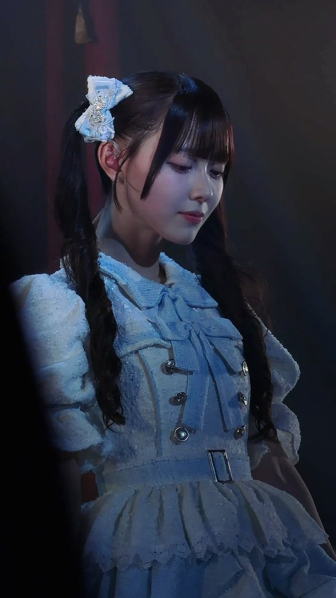 A young woman with braided hair and a decorative white bow is captured on stage wearing an elaborate light blue ruffled dress with button details, her gaze cast modestly downward—embodying the graceful "High Mountain Nadeshiko" referenced in the post about Takane no Nadeshiko and Momo Nan ni Okurimomo.
