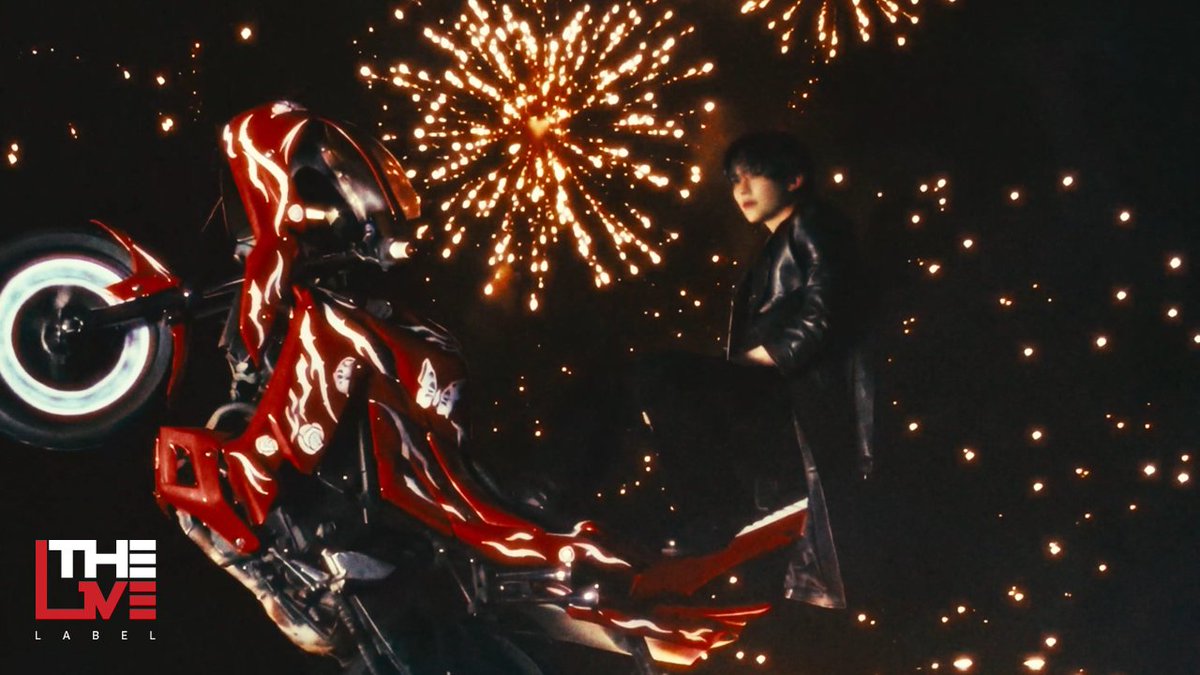 A promotional photo for THE L:LABEL featuring a young man in a long black coat standing next to a red futuristic motorcycle adorned with flame decals, set against a backdrop of bursting fireworks.