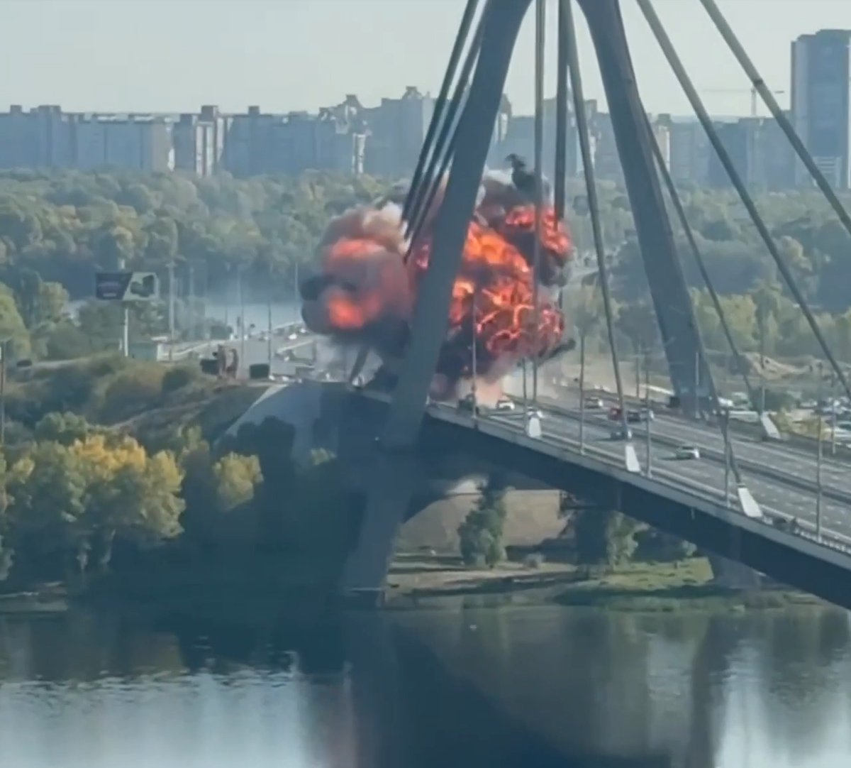 A dramatic explosion captured on a large cable-stayed bridge over a river, with massive fireballs and smoke billowing from the bridge deck while city buildings are visible in the background.