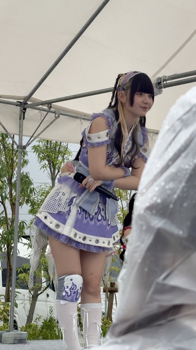 A young woman in a purple and white idol-style performance outfit stands under a tent, holding a microphone.