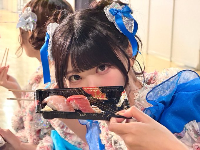 A young woman with dark hair styled in twin buns with blue and white ribbons holds a small black tray of sushi playfully in front of her face, wearing a blue and white frilly outfit.
