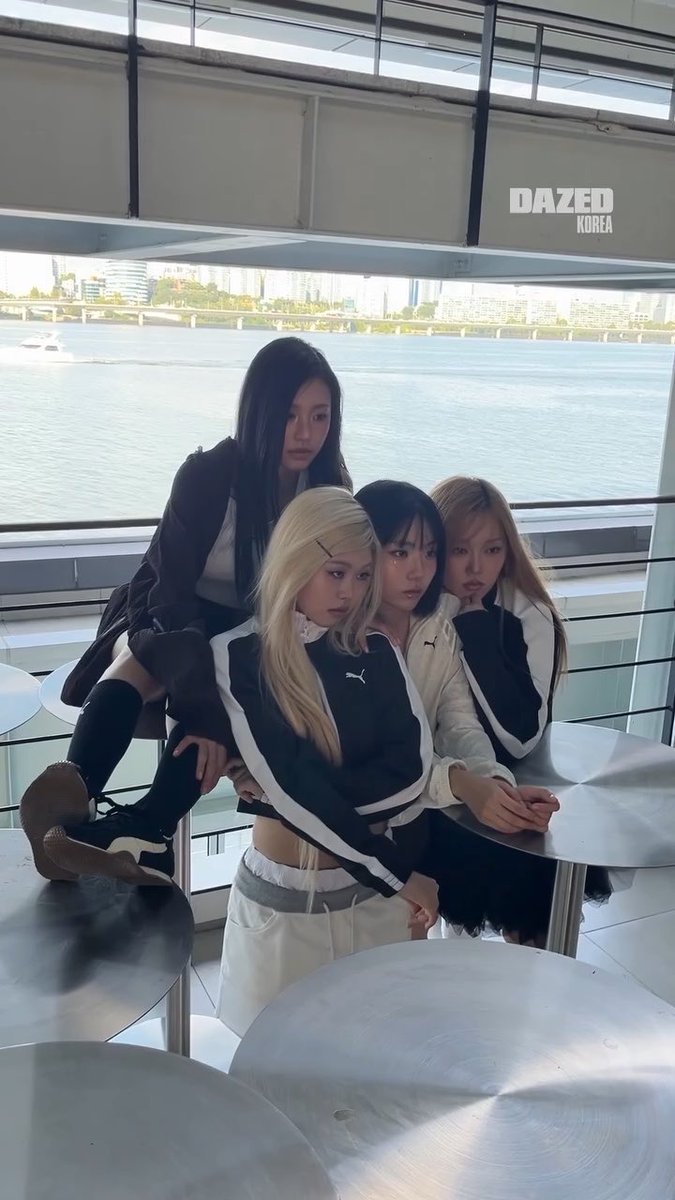 Four women, appearing to be K-pop idols, pose together in a modern space with large windows overlooking a river and city, with 'DAZED KOREA' text visible; they wear stylish black and white outfits.