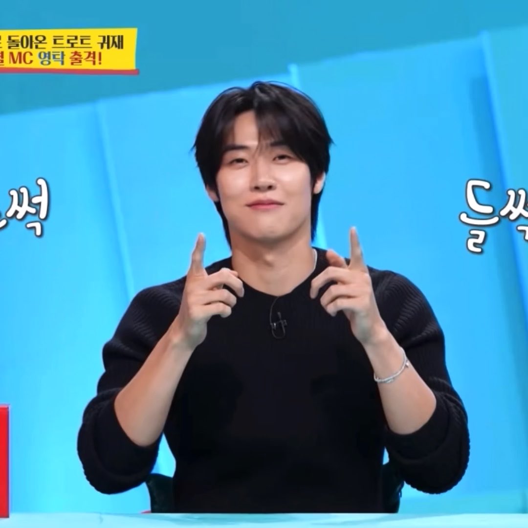 A young man with dark hair smiles and points both index fingers upward on a TV set with Korean text including 'MC 명탁 출격!' visible in the corner.