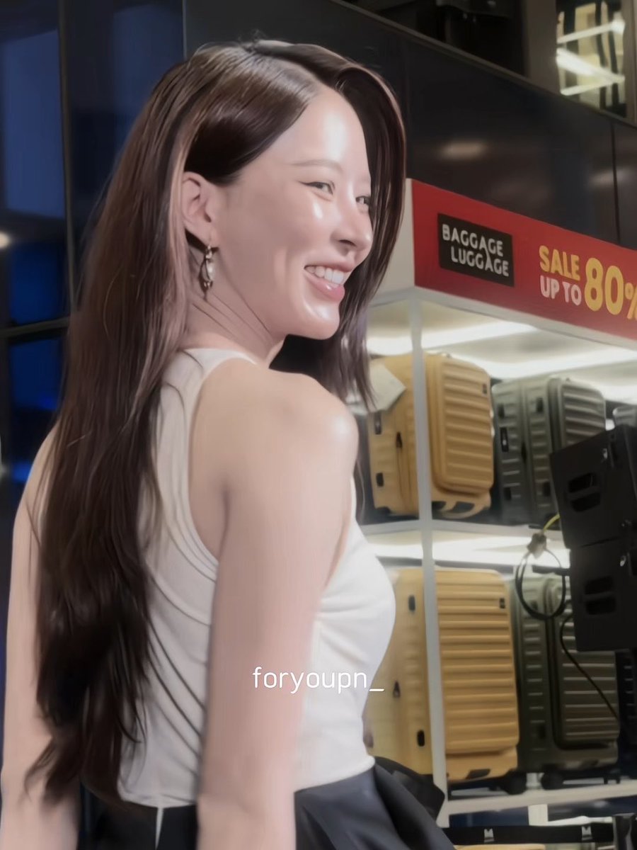 A smiling young woman with long brown hair wearing a white tank top stands in what appears to be a store, with a 'BAGGAGE LUGGAGE SALE UP TO 80%' sign and yellow suitcases visible behind her, watermarked 'foryoupn_'.