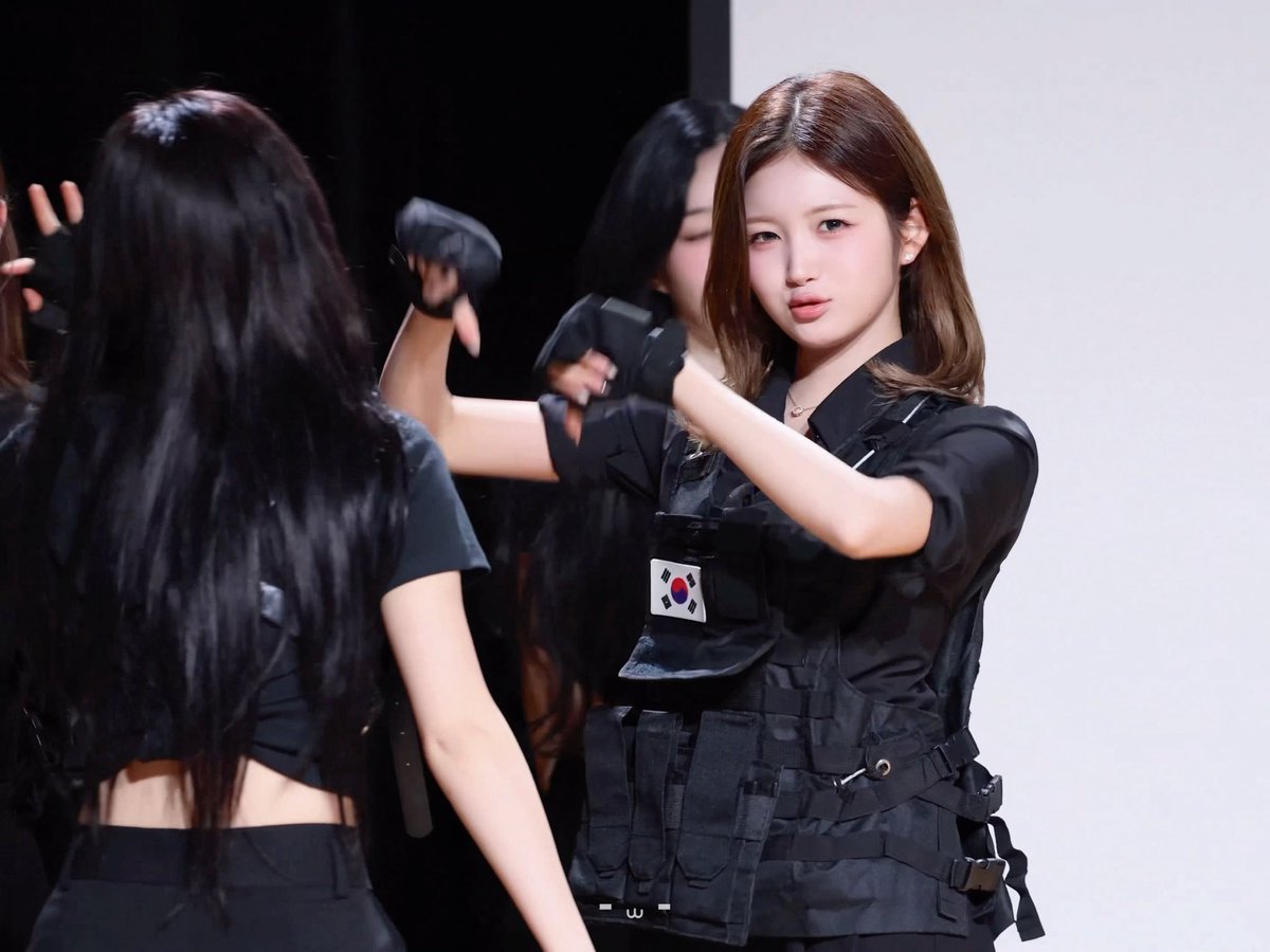 A young woman with brown hair wears a black tactical vest featuring a South Korean flag patch, while other performers in black outfits with gloves are visible in the background, likely rehearsing a choreography. The image relates to the post as it appears to be a behind-the-scenes shot of TripleS members, including potentially Jung Hayeon, during preparation for a performance.