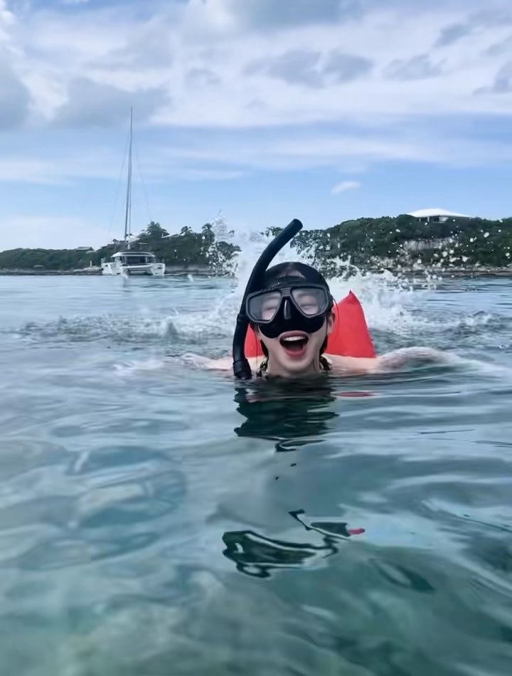 A snorkel mask-wearing KARINA smiles playfully at the camera while swimming in clear tropical waters, with a catamaran and lush hillside visible in the background. The caption "Onii-san, your swimming is so cute and adorable #aespa #KARINA #카리나 #คาริน่า" matches the cheerful, adorable snorkeling moment captured in the image.