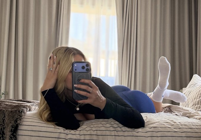 A woman with blonde hair lounges on a striped bed, taking a mirror selfie with her phone while wearing white crew socks and blue leggings, playfully obscuring her face. The image ties to the caption "bark for my gym socks" by showcasing the simple white socks she's asking for attention on.