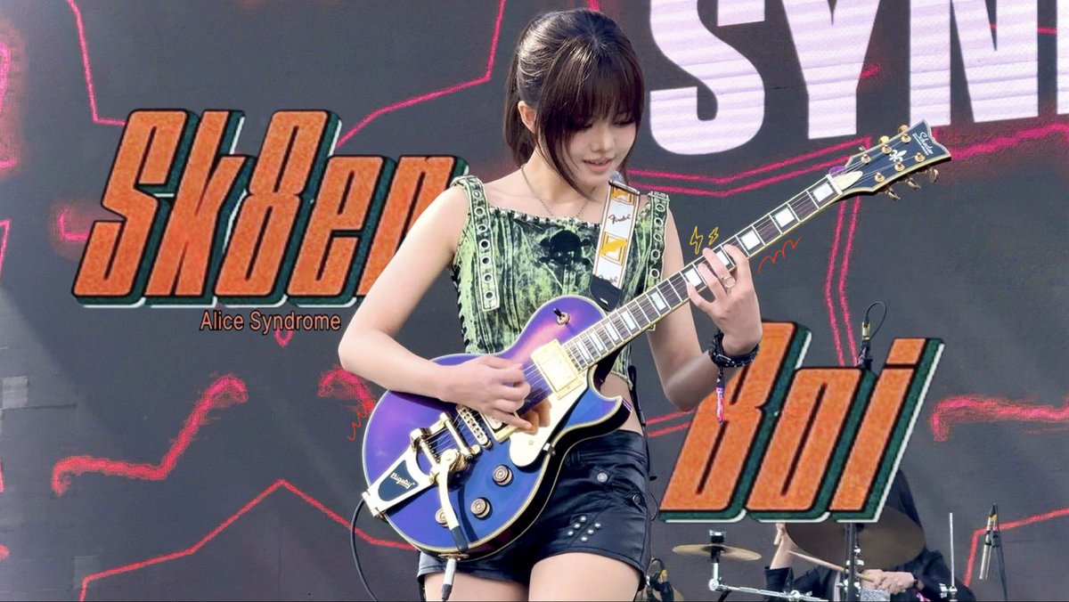 A guitarist playing a purple electric guitar on stage, wearing a green patterned top, with a large screen in the background displaying the words "SYN" and "Alice Syndrome".
