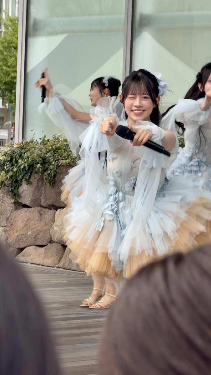 A young performer in a flowing white and peach ruffled dress smiles while holding a microphone at an outdoor stage, with another performer partially visible behind her.