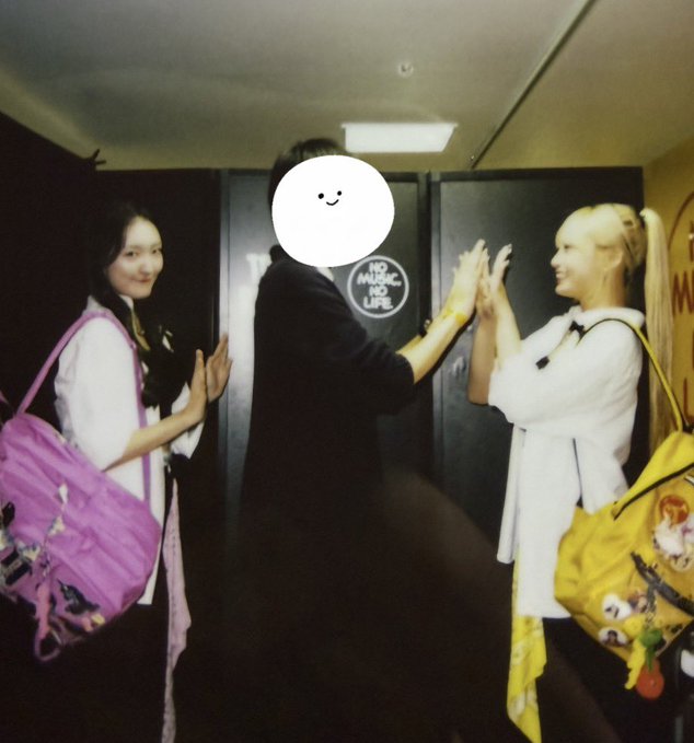Two smiling women high-fiving a third person (face covered by a smiley emoji sticker) in a hallway; one woman carries a purple patterned bag and the other a yellow bag with character designs, with 'NO MUSIC NO LIFE' visible on a door.