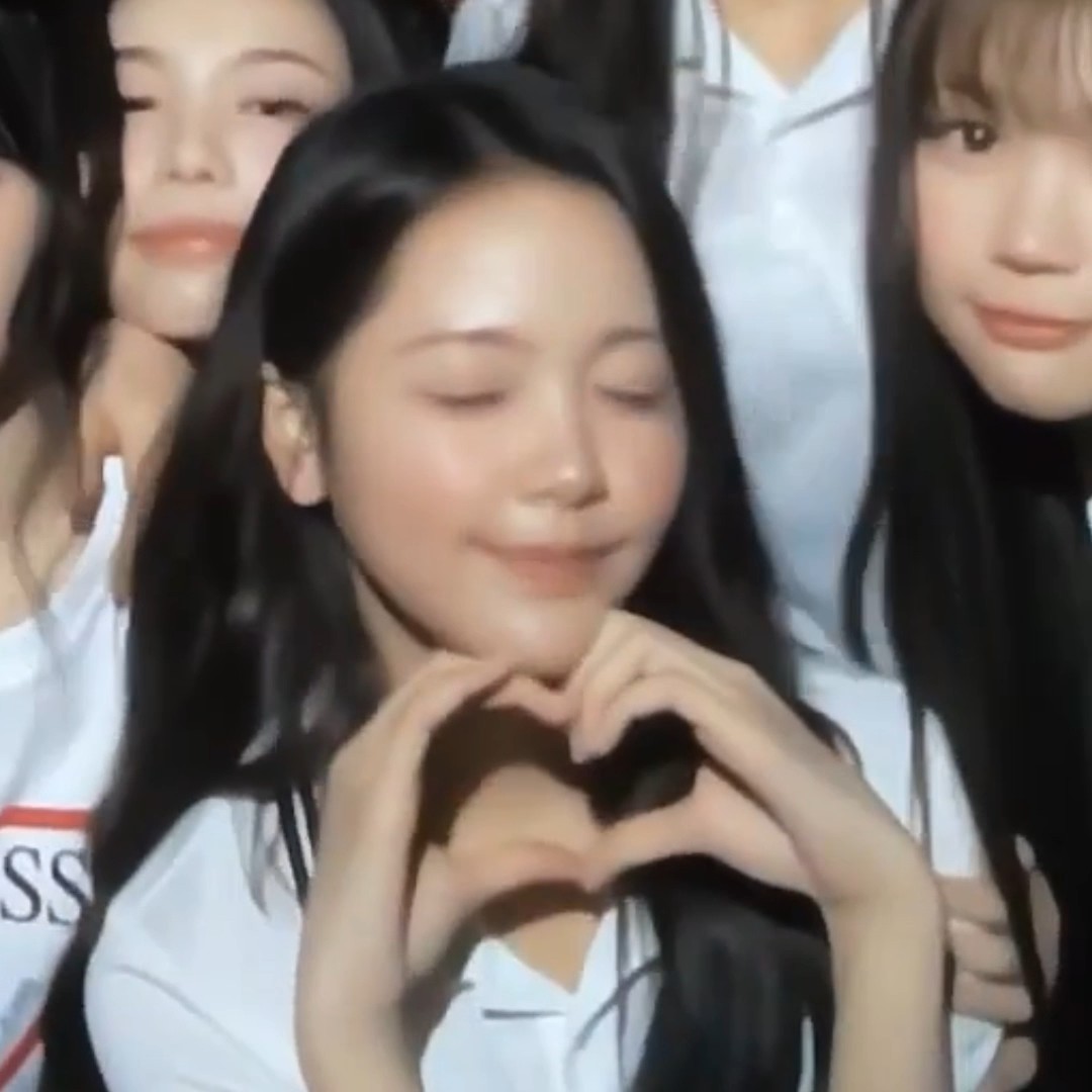 A young woman with long black hair making a heart shape with her hands, eyes closed, with two other women visible behind her.