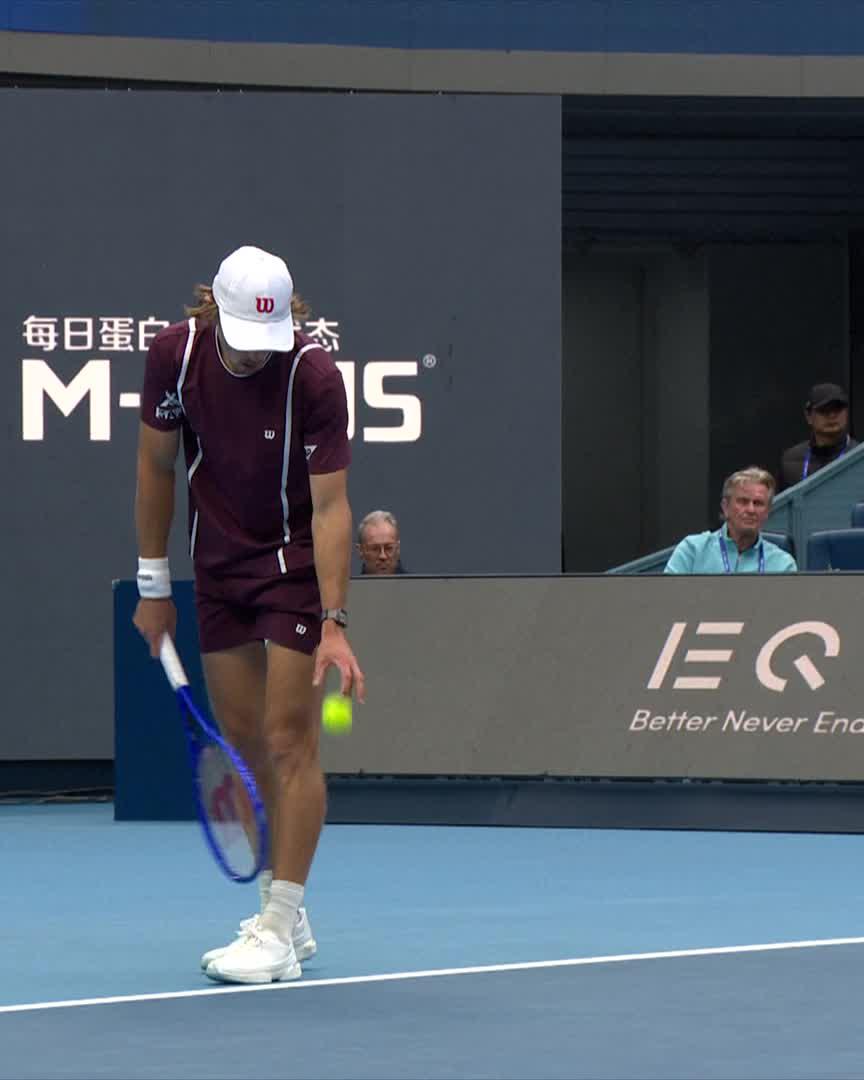 Alex de Minaur, wearing a maroon Wilson kit and white cap, prepares to bounce the tennis ball before serving during his match at the China Open, with "MEG" and Chinese characters visible on the backdrop. The image captures his focused pre-serve routine following his solid three-set victory over Rublev in Beijing.