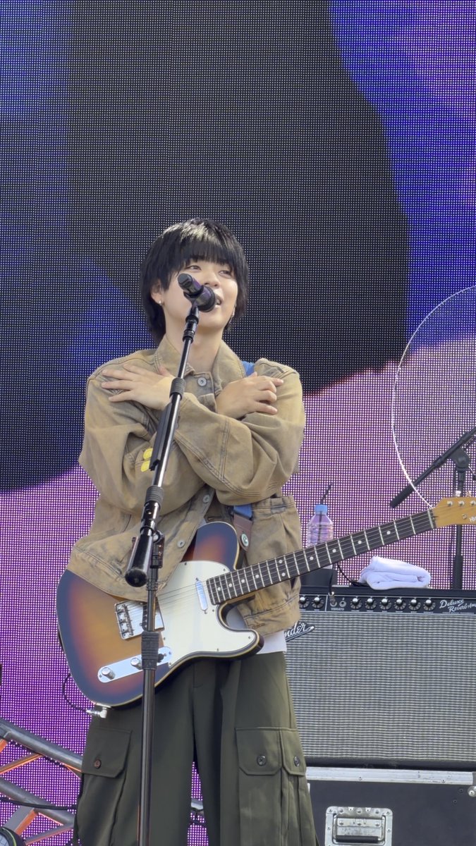 A person with short black hair stands on stage with arms crossed, holding a sunburst Telecaster-style electric guitar, positioned in front of a microphone and amplifier.