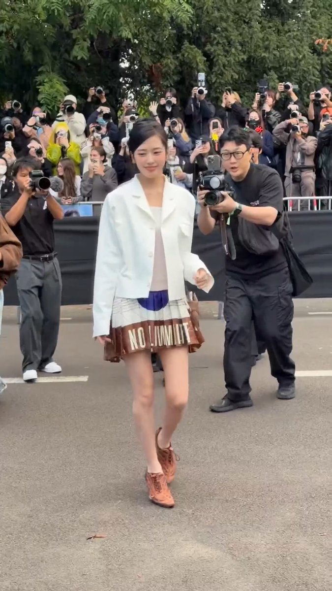 A woman in a white blazer and a short skirt walks down a street surrounded by photographers and videographers.