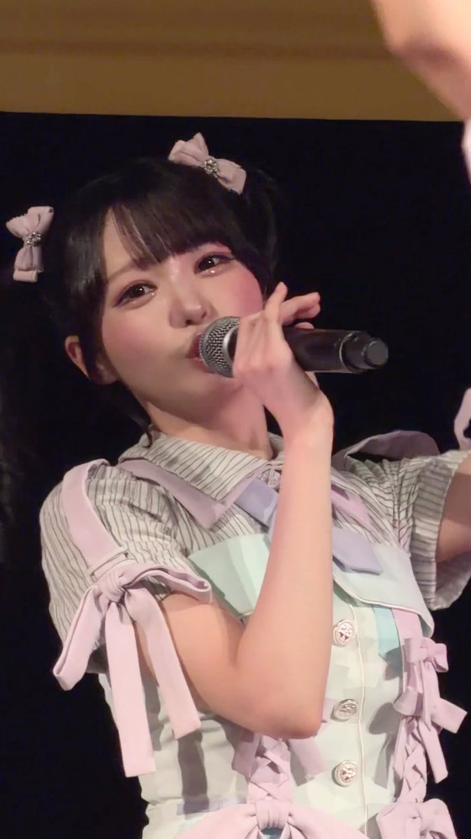 A young Japanese idol performer with pink hair bows sings into a microphone while wearing a pink and white ruffled outfit on stage.