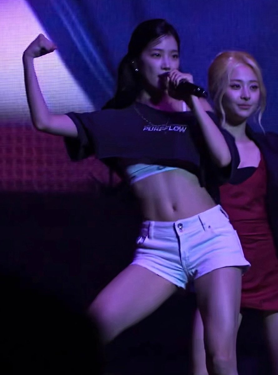 A female performer on stage holds a microphone while flexing her arm, wearing a black cropped t-shirt reading "PURE FLOW" and white denim shorts. The hashtags identify her as Kazuha, a member of the K-pop group LE SSERAFIM, performing during one of the group's concerts.
