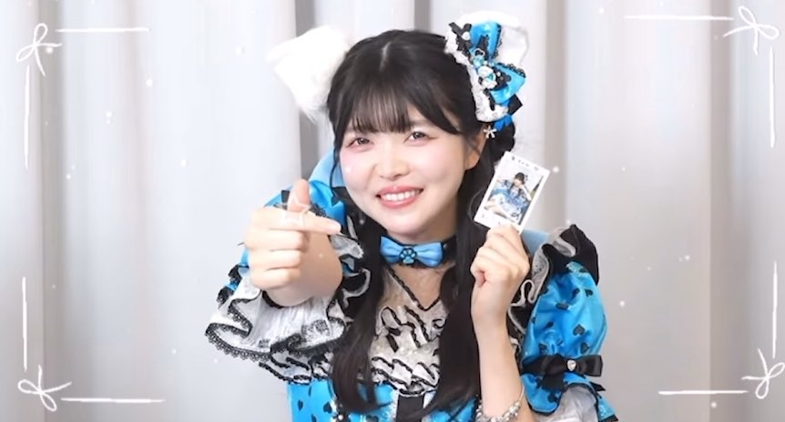 A smiling young East Asian woman wearing a blue frilly outfit with cat-ear accessories, pointing at the camera while holding a photo card in her other hand.