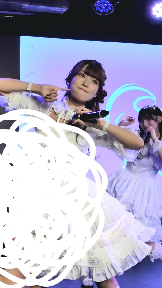 Two women in frilly white dresses performing on a lit stage, one pointing at her cheek while holding a microphone.