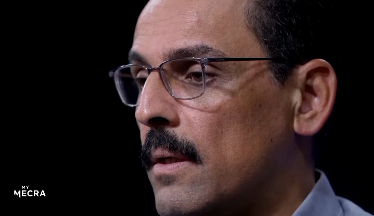 A close-up profile shot of a man with glasses and a mustache speaking, with 'MY MECRA' logo in the bottom left corner against a dark background.