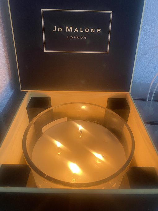 An open black box displays "JO MALONE LONDON" on the inside lid, illuminated by a lit candle within a glass holder.