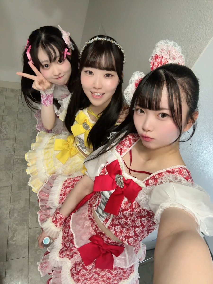 A selfie of three young women posing together, dressed in elaborate frilly idol-style costumes in pink, yellow, and red with large bows and lace headpieces.